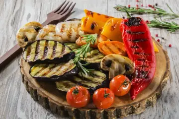 Grillowane warzywa: bakłażan, papryka, pieczarki, pomidorki i marchewka. Idealne dodatki do grilla, podane na drewnianej desce.